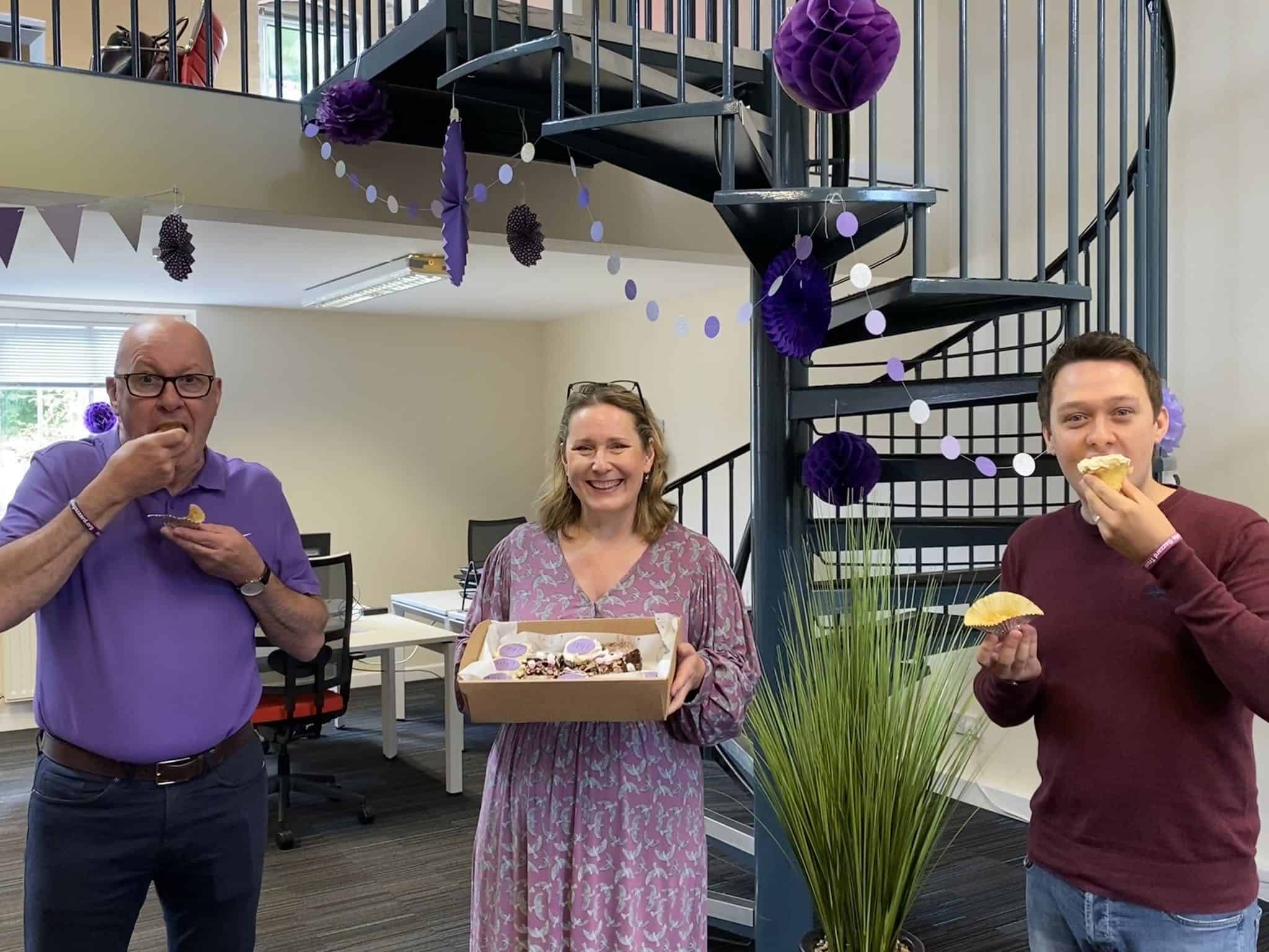 Enjoying a celebration with cake and cupcakes in an office decorated with purple paper lanterns and banners, featuring colleagues sharing joyful moments at work.