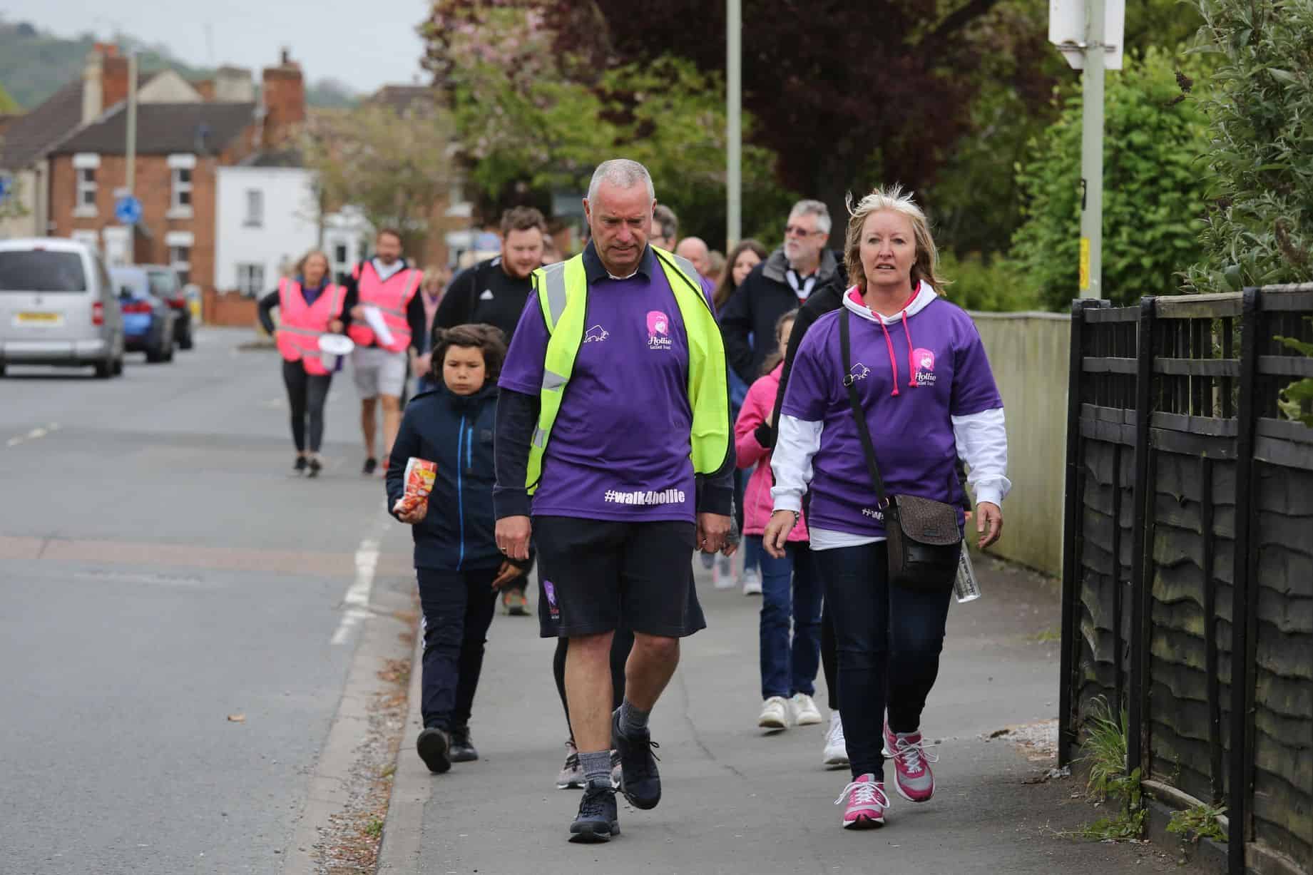 Engaging community walk in a neighbourhood with people wearing purple #walk4hollie shirts for charity awareness and support. Group of diverse participants walking together on a peaceful street.