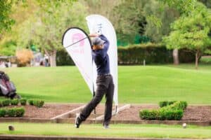 1. Male golfer swinging club on lush golf course during daytime, surrounded by trees and promotional flags.