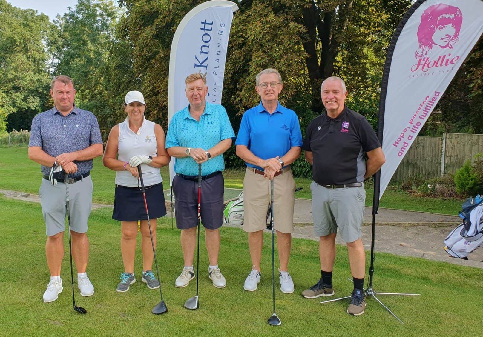 1. Group of six golfers standing on the golf course with flags displaying "Hollie Gazzard Trust" and "CK Knowledge," smiling after a charity golf event.