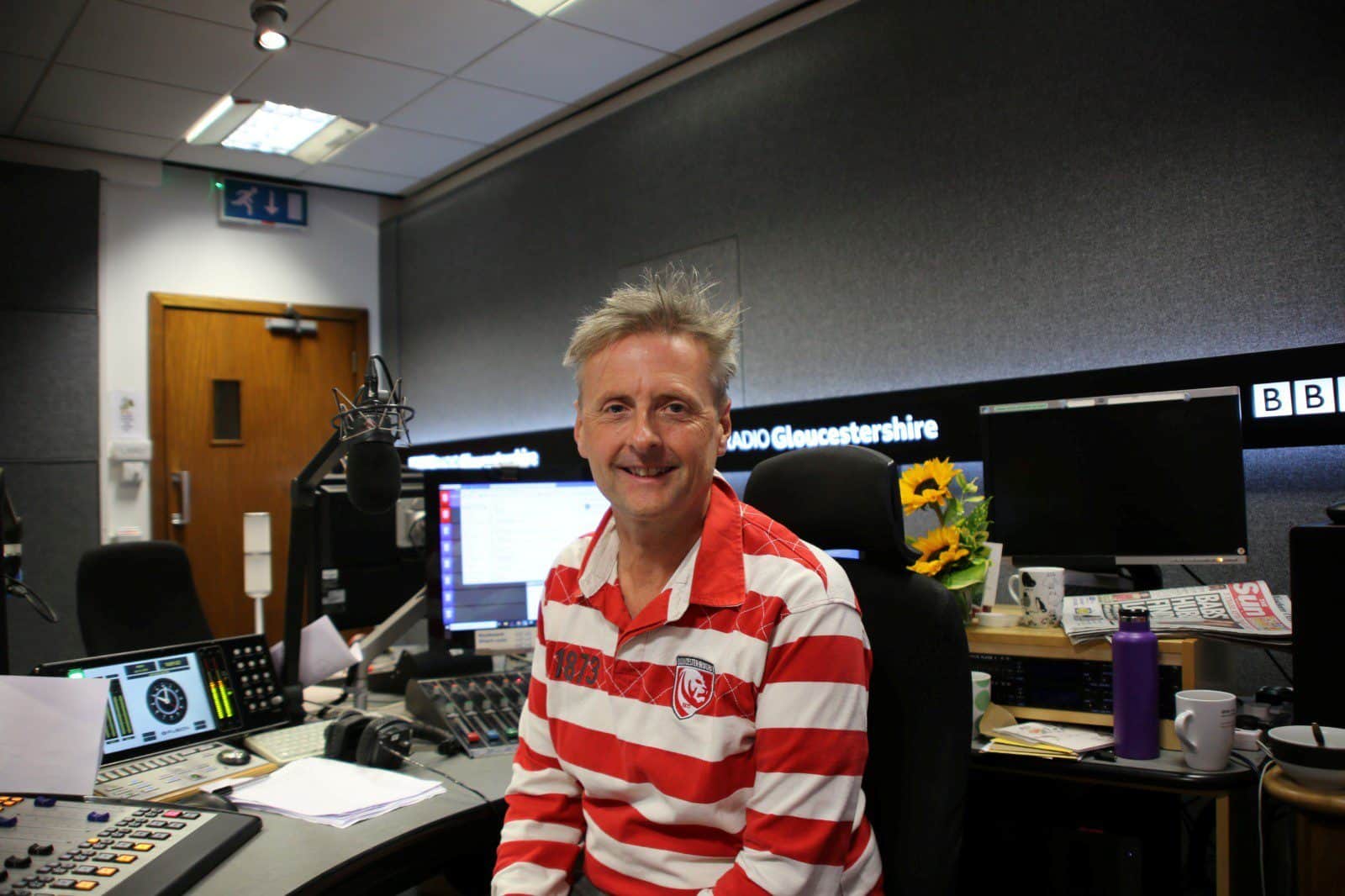 Radio presenter in the BBC Gloucestershire studio with broadcasting equipment, microphones, and monitors for radio production.