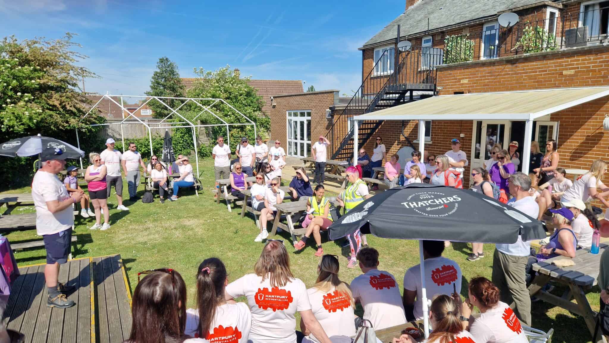 People attending an outdoor community event in a garden, seated on picnic benches under bright sunshine, with speakers and organisers engaging the crowd for social or charity activities.
