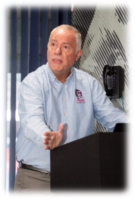 Aged man in a light blue shirt giving a presentation or speech in an office or conference room setting.