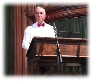 Energetic male performer at a vintage wooden piano during a live musical event, dressed in a white shirt and red bow tie, delivering a captivating performance in a classic setting.