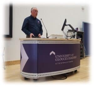 A university lecturer presents at a lectern during a lecture at the University of Gloucestershire, with a computer monitor and microphone setup in a modern lecture theatre.