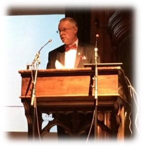 A man in a tuxedo speaking at a wooden podium during a formal event, with microphones and a bright screen in the background.